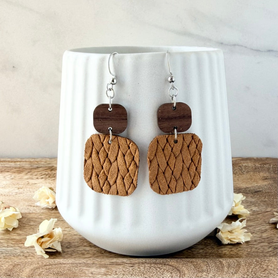 A pair of handmade dangle earrings with dark brown wooden top plates and textured mustard woven geometric drop shapes, hanging from silver fishhook wires. They are displayed on a white ribbed ceramic mug set on a rustic wooden surface with scattered dried flowers.