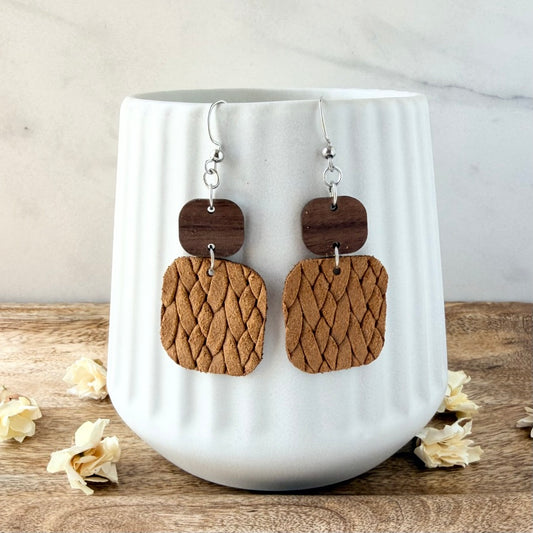 A pair of handmade dangle earrings with dark brown wooden top plates and textured mustard woven geometric drop shapes, hanging from silver fishhook wires. They are displayed on a white ribbed ceramic mug set on a rustic wooden surface with scattered dried flowers.