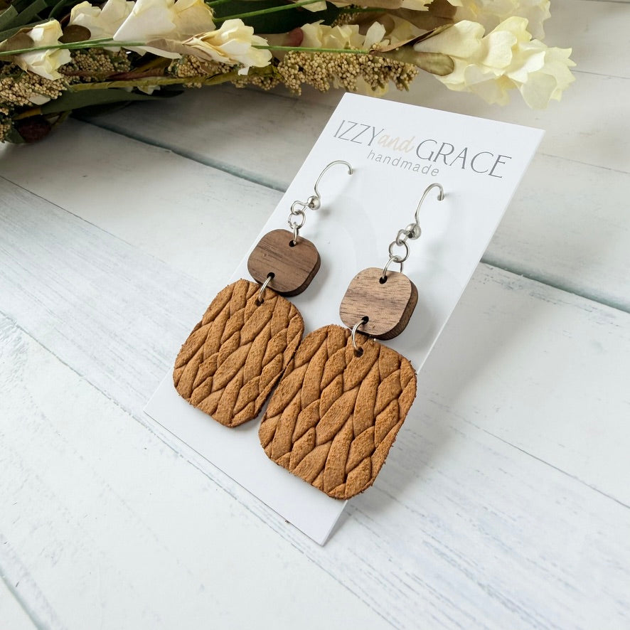 A pair of handmade dangle earrings displayed on a white card. Each earring has a silver fishhook wire, a small square dark wood top, and a larger textured, woven mustard leather rectangular drop.