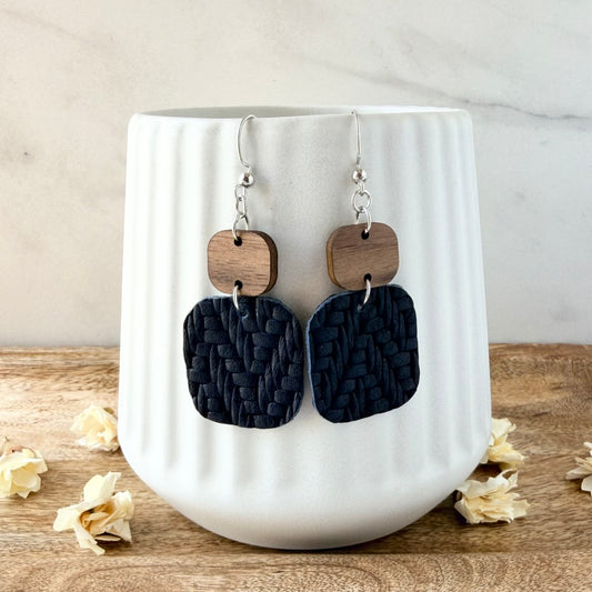 A pair of handmade dangle earrings with dark navy woven leather square drops and natural wood round connectors, hanging from silver fishhook wires in front of a white ribbed ceramic mug.