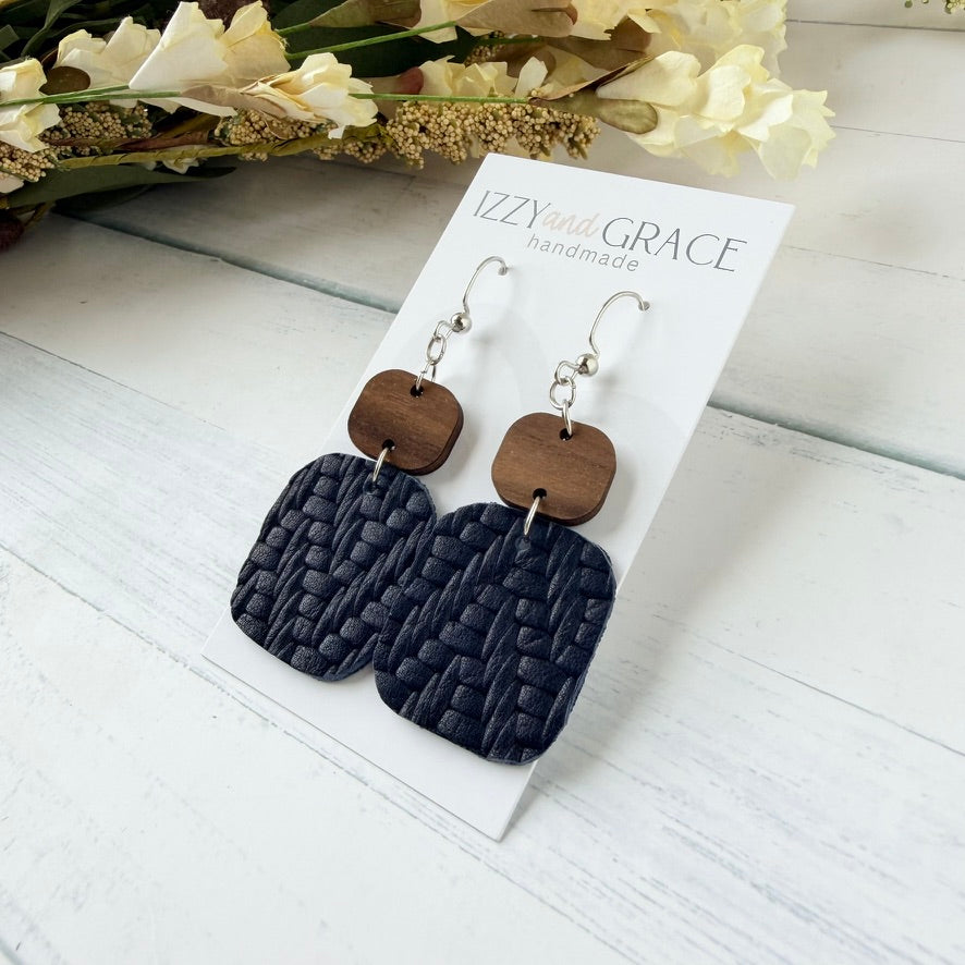 A pair of handmade dangle earrings displayed on a white card labeled 'IZZY with GRACE handmade.' The earrings have silver-tone hooks, brown wooden top, and larger textured dark navy woven rectangular drops.