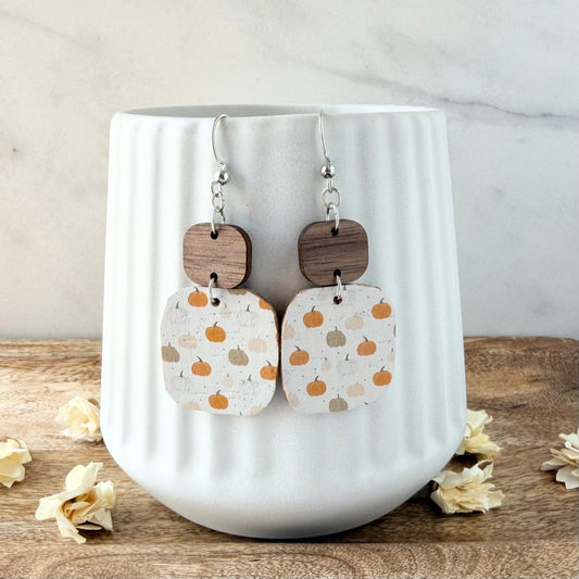 A pair of dangle earrings displayed on a white ribbed ceramic mug. The earrings feature silver fishhook wires, round dark brown wood charms, and square cream-colored charms printed with a pattern of orange and beige pumpkins.