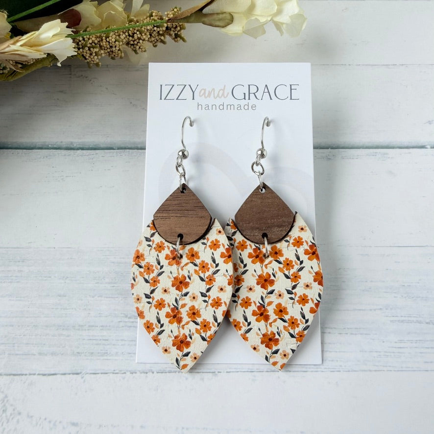 A pair of handmade dangle earrings displayed on a white card labeled 'IZZY and GRACE handmade.' The earrings have silver fishhook wires, brown wooden teardrop tops, and cream leather bottoms with an orange floral print.