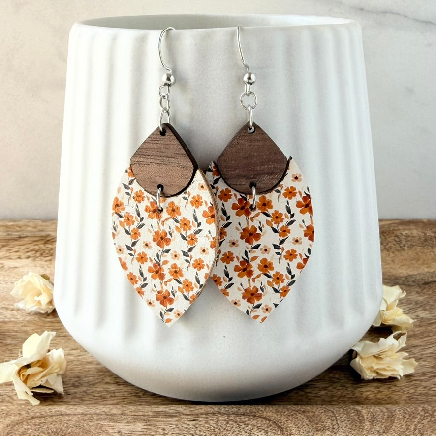 A pair of dangling earrings with silver fishhook wires and teardrop-shaped wooden tops connected to larger petal-shaped dangles printed with an orange floral pattern on a cream background. The earrings are displayed on a white ribbed ceramic mug set on a wooden surface with dried flowers.