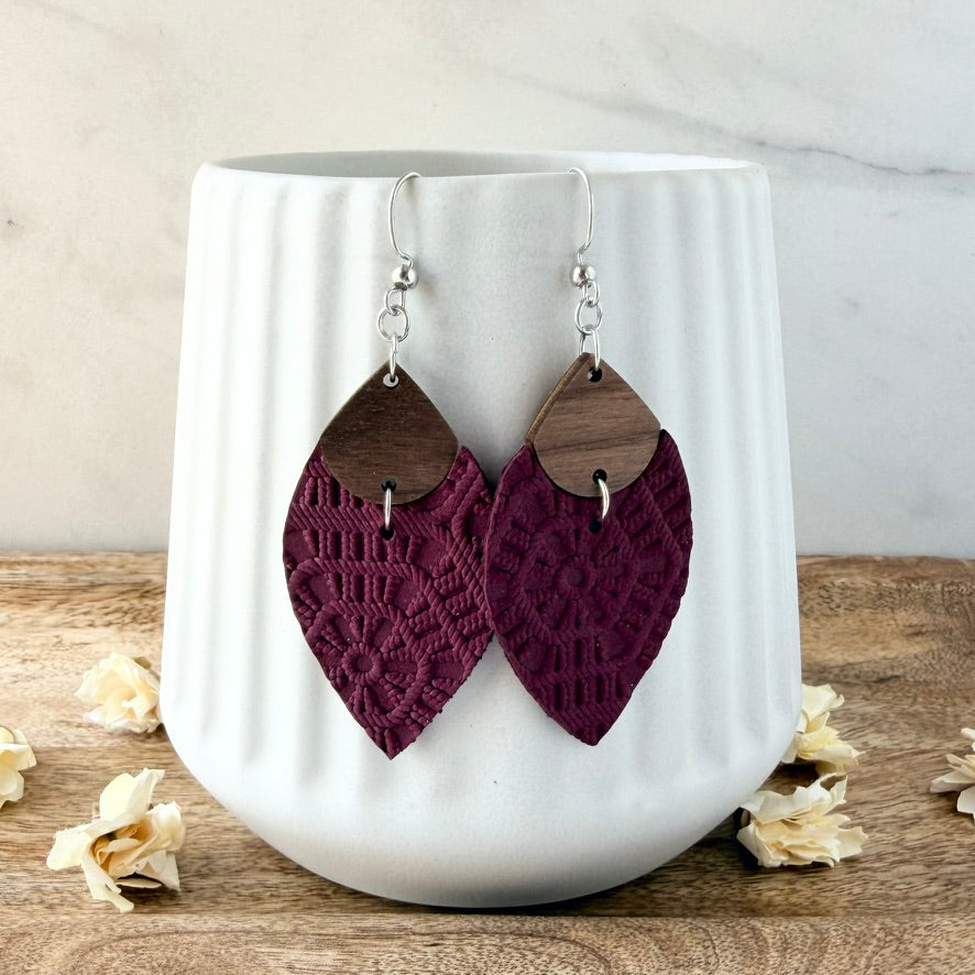 A pair of handmade dangle earrings with dark brown wooden teardrop-shaped tops and deep burgundy lace-textured lower halves, hanging from silver fishhook wires in front of a white ribbed ceramic vase.