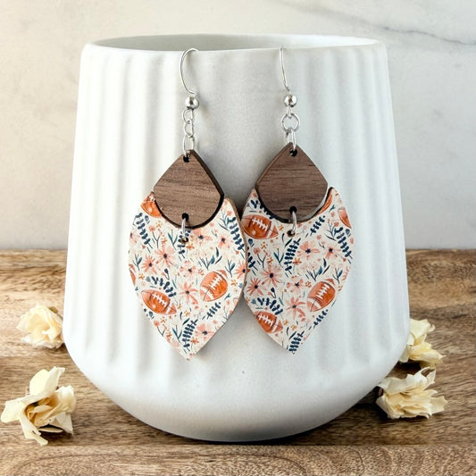 A pair of dangling earrings with floral and football patterns on a white ceramic mug, set on a wooden surface with dried flowers.