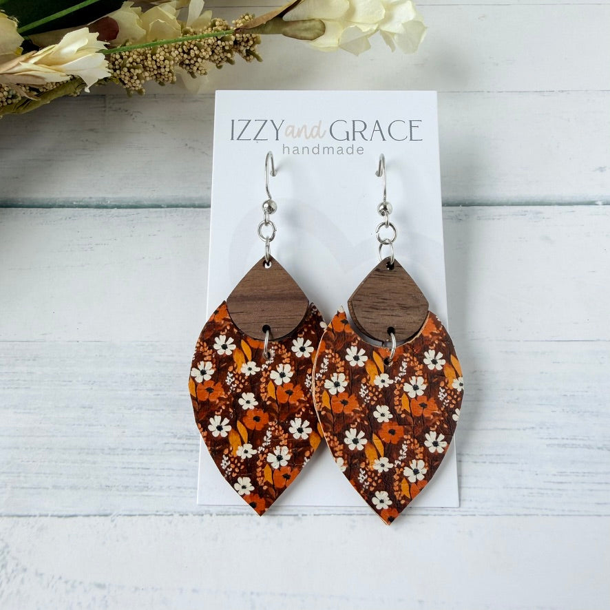 A pair of dangling earrings displayed on a white card labeled 'IZZYand GRACE handmade.' The earrings have silver fishhook wires and teardrop-shaped wooden tops with a floral pattern in orange, brown, and white.