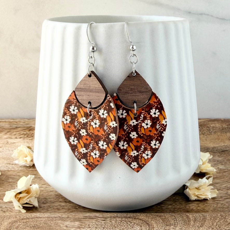 A pair of petal-shaped dangle earrings with a brown wood top half and a patterned lower half in orange, brown, and white floral motifs, hanging from silver fishhook wires in front of a white ribbed ceramic mug.