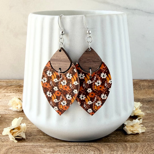 A pair of petal-shaped dangle earrings with a brown wood top half and a patterned lower half in orange, brown, and white floral motifs, hanging from silver fishhook wires in front of a white ribbed ceramic mug.