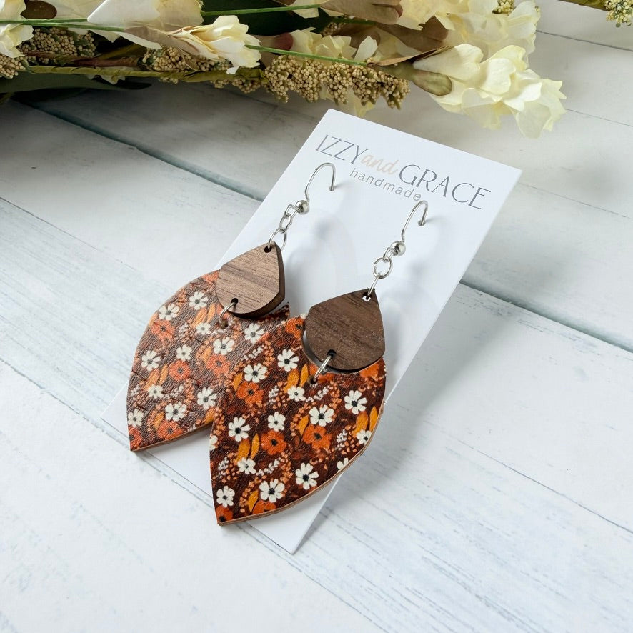 A pair of handmade dangle earrings displayed on a white card labeled 'IZZY and GRACE handmade.' The earrings have silver fishhook wires and teardrop-shaped wooden tops, and leather bottoms with a white and orange floral pattern on a brown background.