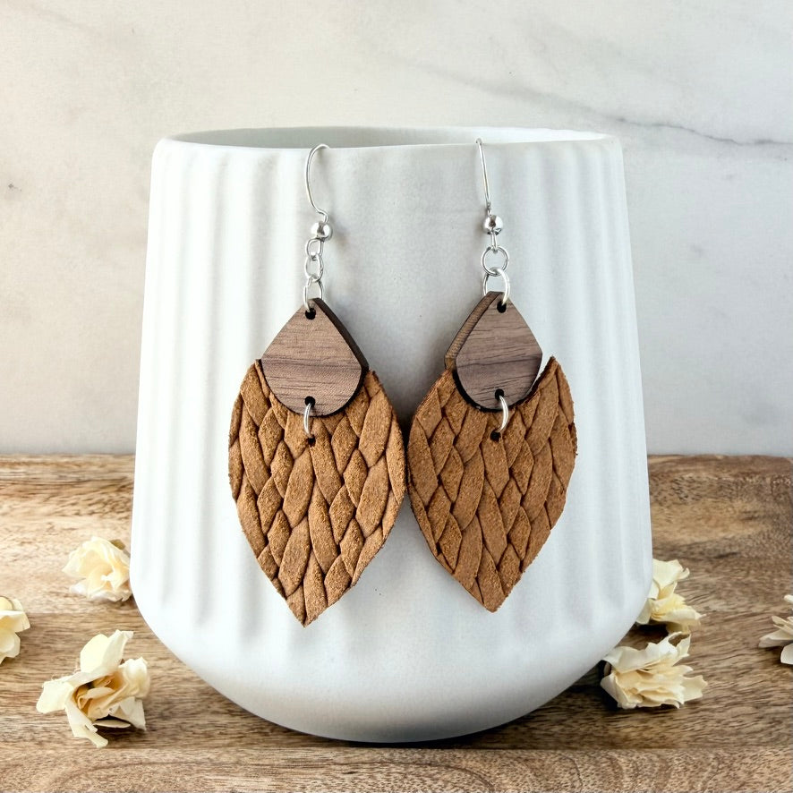 A pair of handmade dangle earrings with textured mustard leather and dark wood details, hanging from silver hooks on a white ceramic mug.