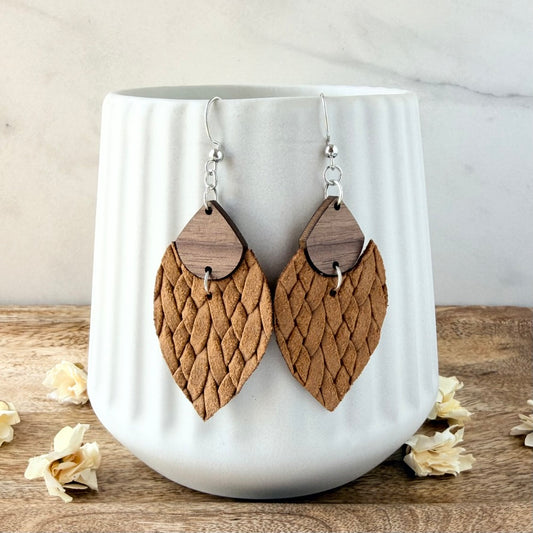A pair of handmade dangle earrings with textured mustard leather and dark wood details, hanging from silver hooks on a white ceramic mug.