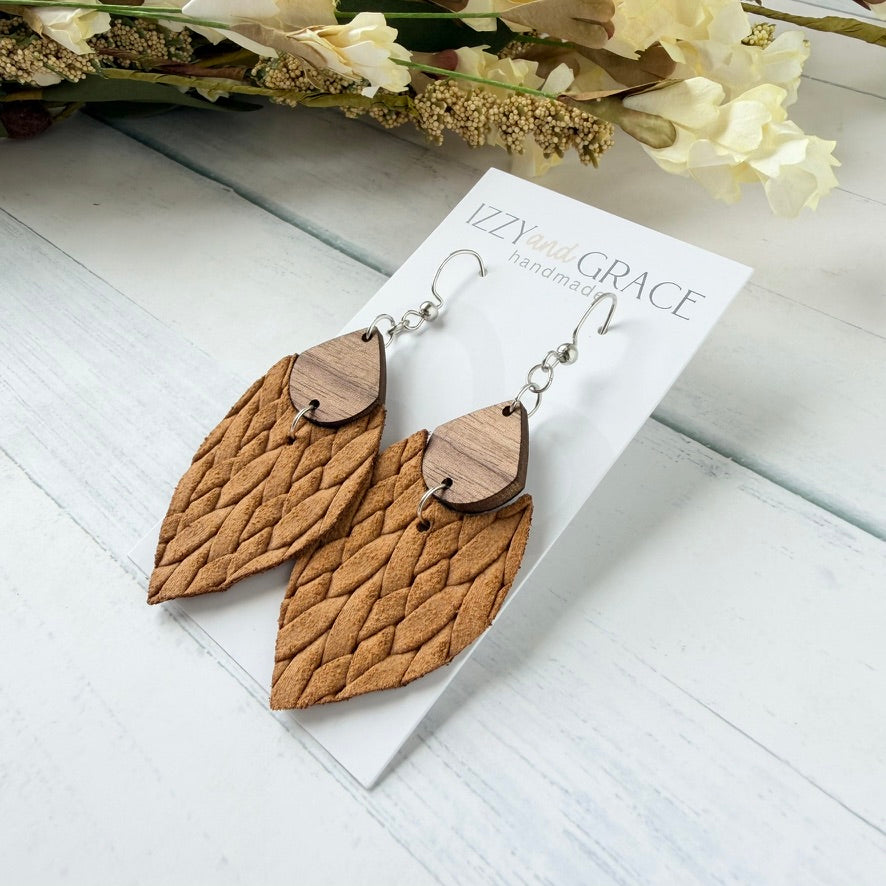 A pair of handmade dangle earrings with textured brown woven leather petal-shaped drops and small wooden teardrops, displayed on a white card labeled 'Izzy and Grace handmade' on a white wood background with dried flowers.