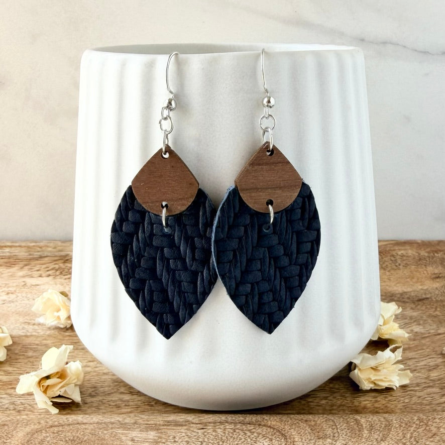 A pair of large petal-shaped dangle earrings with dark navy woven leather lower halves and natural wood top sections, hanging in front of a white ribbed ceramic vase on a wooden surface.