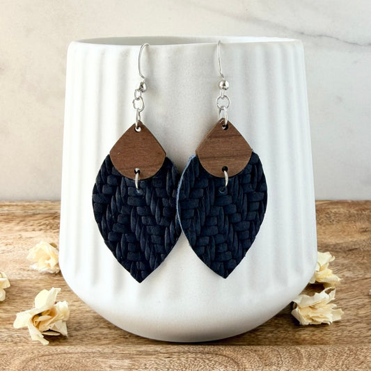 A pair of large petal-shaped dangle earrings with dark navy woven leather lower halves and natural wood top sections, hanging in front of a white ribbed ceramic vase on a wooden surface.