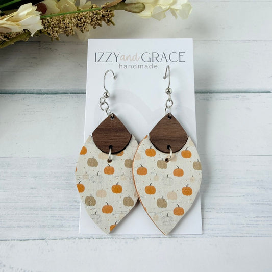 petal-shaped dangle earrings with a cream leather body printed with orange and tan pumpkins, a dark brown wooden top, and silver fishhook wires, displayed on a white card labeled 'IZZY and GRACE handmade' on a white wood surface with dried flowers in the corner.