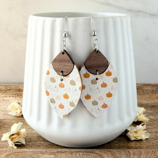 A pair of dangling earrings with silver fishhook wires and teardrop-shaped top. The lower section is printed with small orange and tan pumpkins, displayed in front of a white ribbed ceramic mug on a wooden surface.